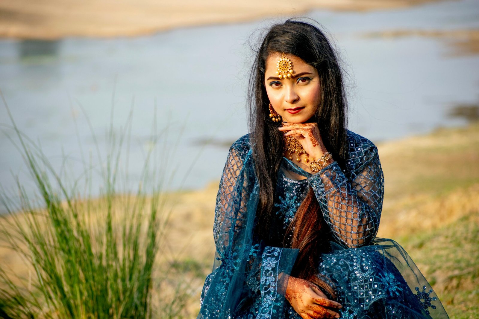 A woman in blue traditional Indian attire with jewelry sitting gracefully by the river.